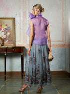 A woman stands indoors facing away, wearing a sheer lavender top and the Social Queen 3D Floral Embroidered Silk Skirt, gold heels, and holding a small woven handbag. A painting and table are visible in the background.