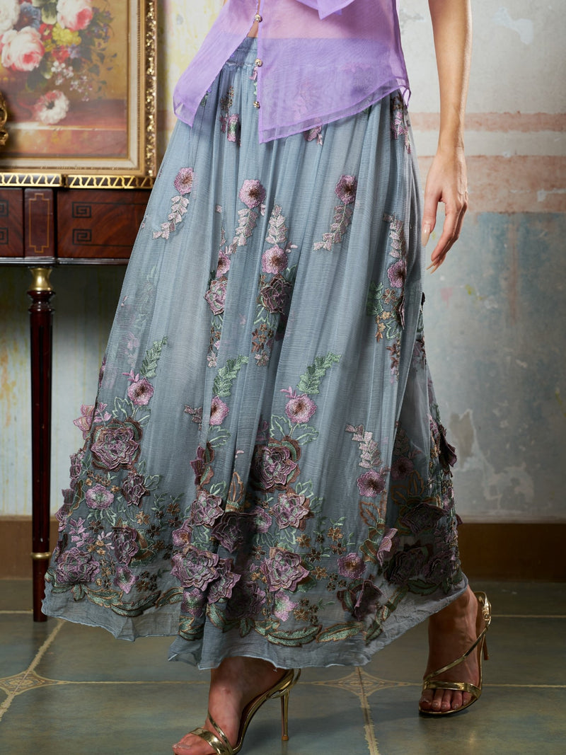 A person stands indoors near a table and framed painting, wearing Social Queen’s 3D Floral Embroidered Silk Skirt with a sheer lavender blouse and heeled sandals.
