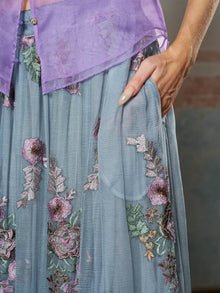 A person with manicured nails rests their hand in the pocket of the Social Queen 3D Floral Embroidered Silk Skirt, a sheer blue midi skirt adorned with handcrafted purple and green floral details.