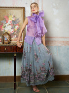 A woman poses by a wooden table, wearing Social Queen’s 3D Floral Embroidered Silk Skirt and a sheer lavender bow blouse, with floral artwork and a framed photo set against a textured wall.