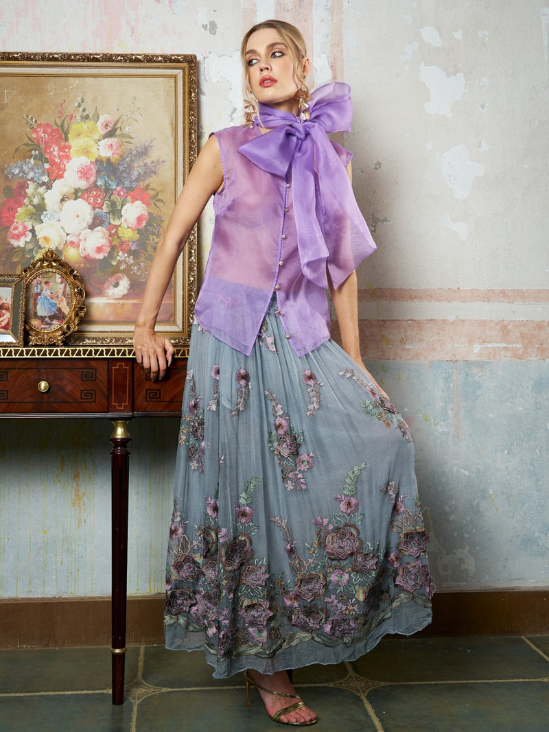 A woman poses by a wooden table, wearing Social Queen’s 3D Floral Embroidered Silk Skirt and a sheer lavender bow blouse, with floral artwork and a framed photo set against a textured wall.