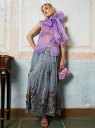 A woman models the Social Queen 3D Floral Embroidered Silk Skirt with a sleeveless purple top, sandals, and a matching clutch, standing against a textured wall.