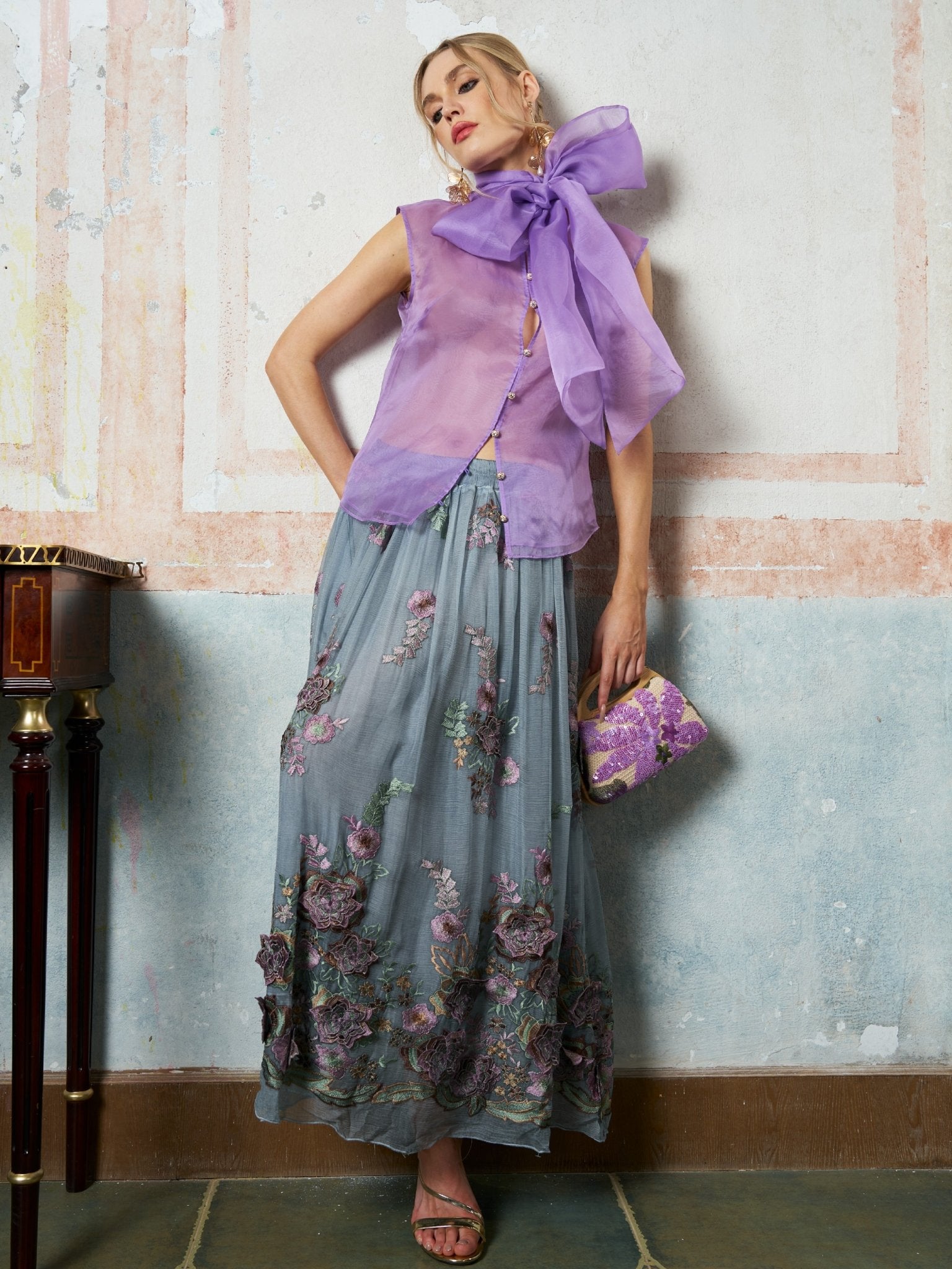 A woman models the Social Queen 3D Floral Embroidered Silk Skirt with a sleeveless purple top, sandals, and a matching clutch, standing against a textured wall.
