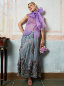 A woman models the Social Queen 3D Floral Embroidered Silk Skirt with a sleeveless purple top, sandals, and a matching clutch, standing against a textured wall.