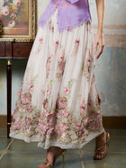 A person wearing Social Queen’s 3D Floral Embroidered Silk Skirt and gold heels stands indoors beside a wooden table and a framed painting.