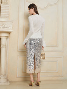 A woman stands indoors facing away, wearing the Social Queen 3D Floral Lace Wool-Silk Knit Top with ruffled lace cuffs and a silver sequin skirt. She holds a small bag and wears gold high-heeled sandals.