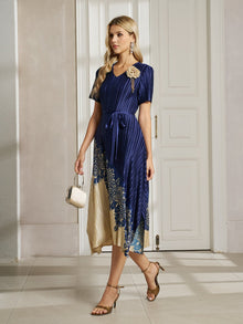 Indoors, a woman wears the Social Queen 3D Floral Plissé Satin Lace-Print Dress in blue and beige, paired with heeled sandals and a small handbag.