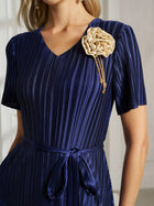 A woman in the Social Queen 3D Floral Plissé Satin Lace-Print Dress, featuring short sleeves, pleats, a fabric belt, and a beige floral appliqué brooch with dangling beads on the chest.