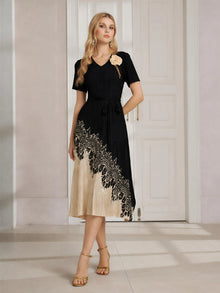 A woman stands indoors in front of white paneled doors, wearing the Social Queen 3D Floral Plissé Satin Lace-Print Dress with a belted waist and gold sandals.