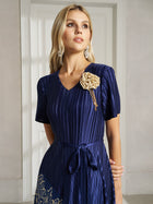 Woman wearing the Social Queen 3D Floral Plissé Satin Lace-Print Dress with a V-neck and short sleeves, styled with large earrings and a beige flower brooch, standing indoors in front of a white door.