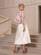 A woman stands sideways on stairs indoors, wearing a patterned long-sleeve top, heeled sandals, and the Social Queen 3D Floral Satin Midi Skirt.
