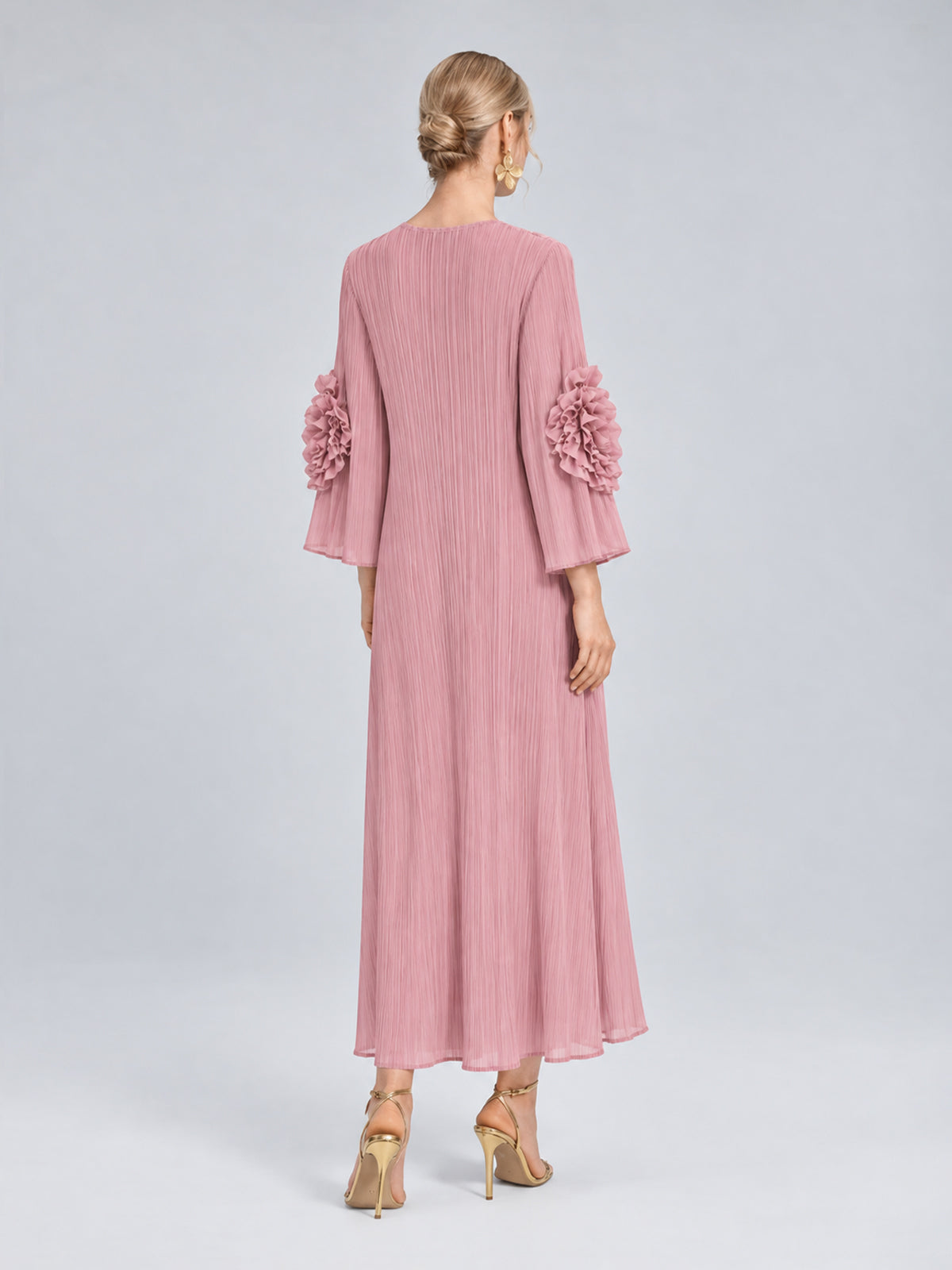 A woman stands with her back to the camera, wearing the Social Queen 3D Floral Pleated Dress—a pink, long-sleeve maxi dress featuring pleats and delicate 3D floral appliqué embellishments on the sleeves—paired with gold high heels.