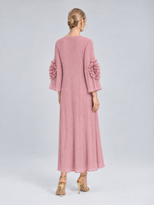 A woman stands with her back to the camera, wearing the Social Queen 3D Floral Pleated Dress—a pink, long-sleeve maxi dress featuring pleats and delicate 3D floral appliqué embellishments on the sleeves—paired with gold high heels.