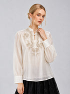 Model is wearing the Social Queen Floral Vine Embroidered Blouse, featuring long sleeves, an ivory hue with delicate floral vine embroidery, and a relaxed straight silhouette. She pairs it with a black skirt and stands against a plain gray background.