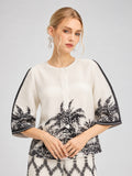 A woman stands against a light background wearing the Social Queen Palm Tree Print Pleated Blouse, featuring a white base with black palm tree print, wide sleeves, and button details, accessorized with dangling earrings.