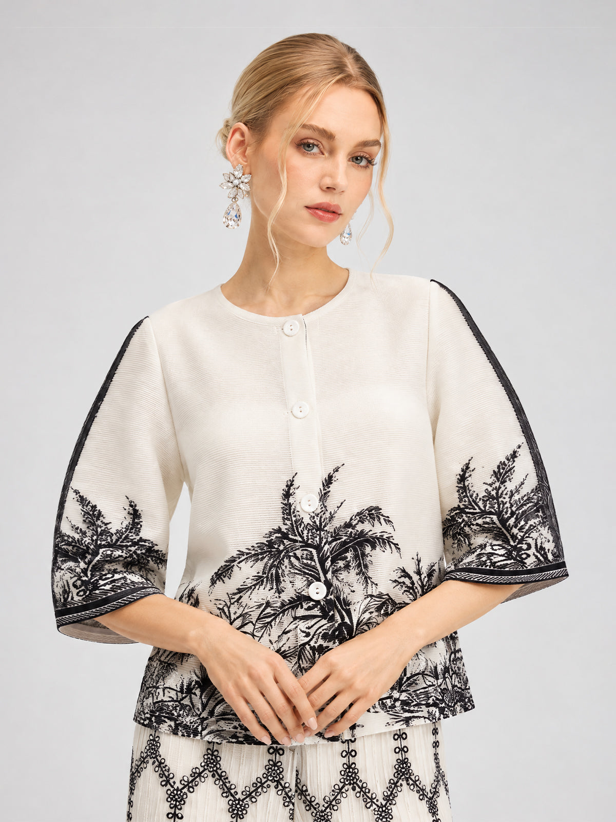 A woman stands against a light background wearing the Social Queen Palm Tree Print Pleated Blouse, featuring a white base with black palm tree print, wide sleeves, and button details, accessorized with dangling earrings.