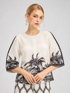 A woman stands against a light background wearing the Social Queen Palm Tree Print Pleated Blouse, featuring a white base with black palm tree print, wide sleeves, and button details, accessorized with dangling earrings.