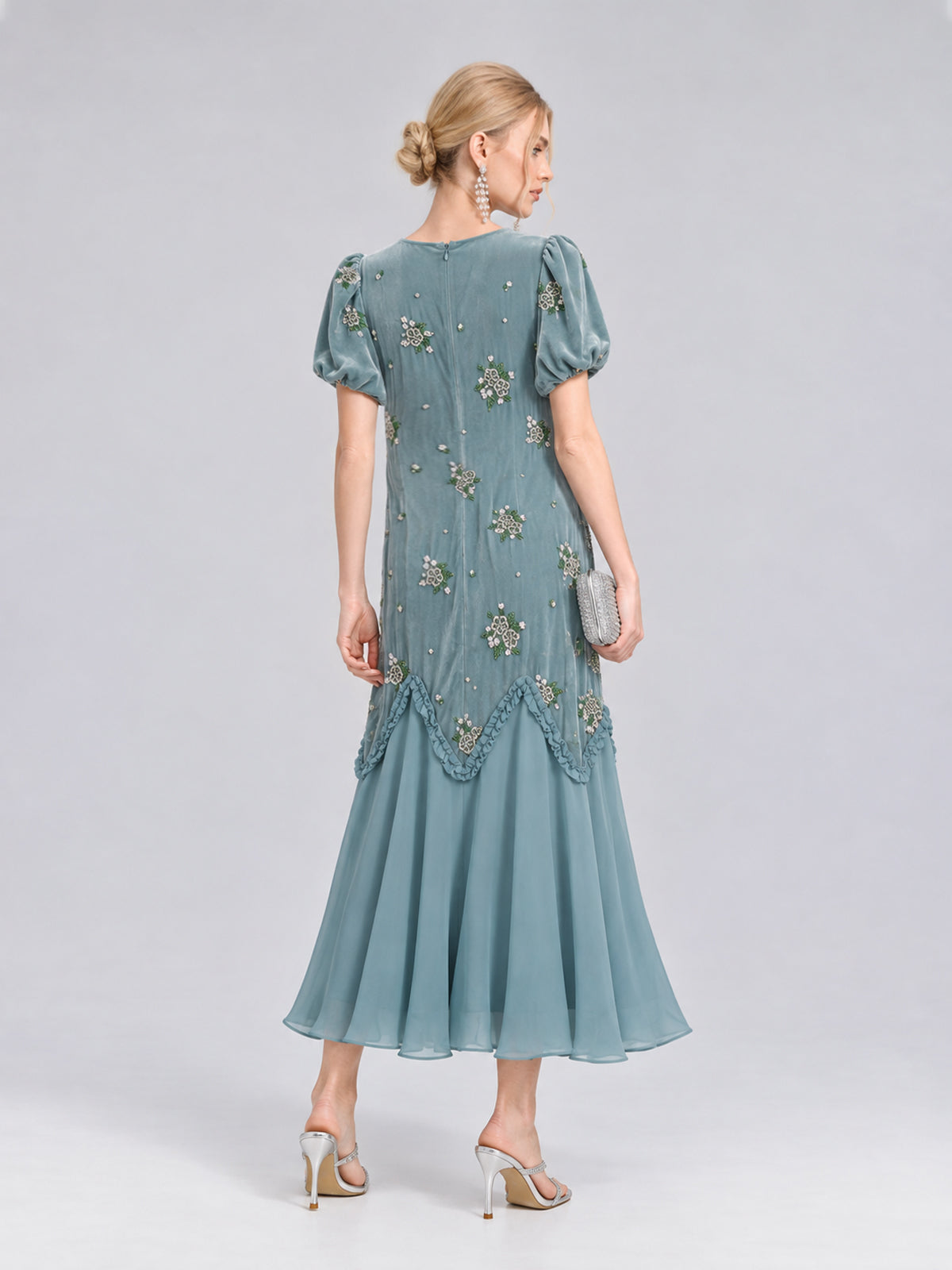 A woman wearing the Beaded Floral Silk Velvet Dress by Social Queen—a teal silk-blend velvet design adorned with beaded floral embroidery, puff sleeves, and a flared hem—holds a clutch and stands in high heels against a plain background, making it the perfect wedding dinner dress.