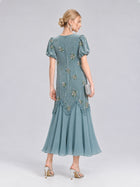 A woman wearing the Beaded Floral Silk Velvet Dress by Social Queen—a teal silk-blend velvet design adorned with beaded floral embroidery, puff sleeves, and a flared hem—holds a clutch and stands in high heels against a plain background, making it the perfect wedding dinner dress.