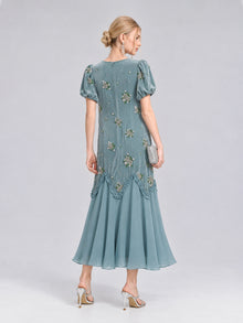 A woman wearing the Beaded Floral Silk Velvet Dress by Social Queen—a teal silk-blend velvet design adorned with beaded floral embroidery, puff sleeves, and a flared hem—holds a clutch and stands in high heels against a plain background, making it the perfect wedding dinner dress.