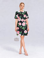 A woman poses against a plain backdrop wearing the Social Queen 3D Peony Floral Shift Dress in black, styled with silver heels, statement earrings, and a petite silver handbag.