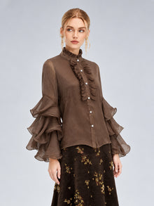 A woman stands against a plain light background wearing the Social Queen Pleated Layered Ruffle Blouse—a sheer brown polyester chiffon top with layered ruffle sleeves and a ruffled front—paired with a dark floral skirt.