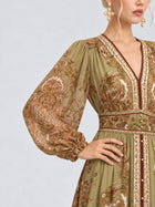 A woman wears the Social Queen Paisley Print Mulberry Silk Dress—an olive green, long-sleeve piece featuring intricate embroidery and a paisley floral print. Paired with dangling earrings and posed with one hand on her hip, she embodies an elegant statement of modern resort wear.