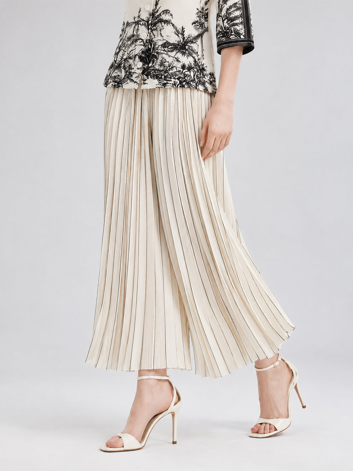 A woman models the Social Queen Striped Pleated Wide-Leg Pants with a black and white patterned top and cream high-heeled sandals, set against a neutral backdrop.