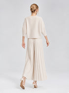 A person stands on a white background, facing away, wearing the Social Queen 3D Floral Pleated Two-Piece Set—featuring a cream-white loose top and pleated wide-leg trousers—paired with high-heeled shoes.