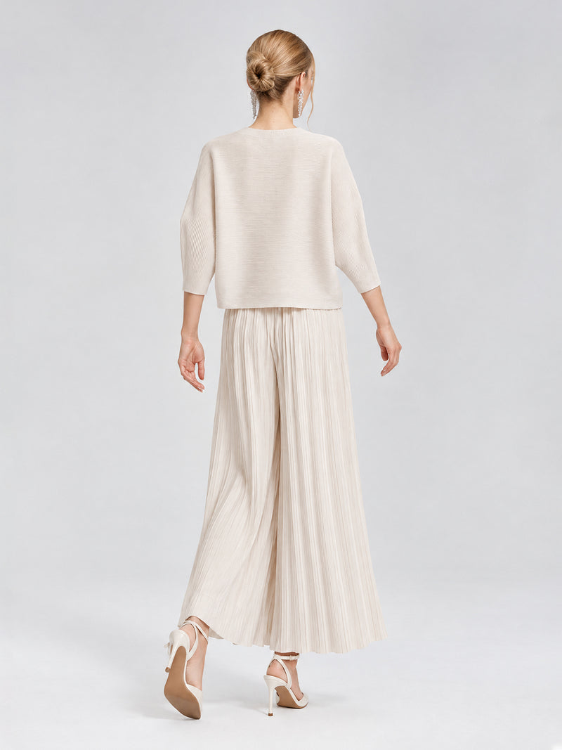 A person stands on a white background, facing away, wearing the Social Queen 3D Floral Pleated Two-Piece Set—featuring a cream-white loose top and pleated wide-leg trousers—paired with high-heeled shoes.