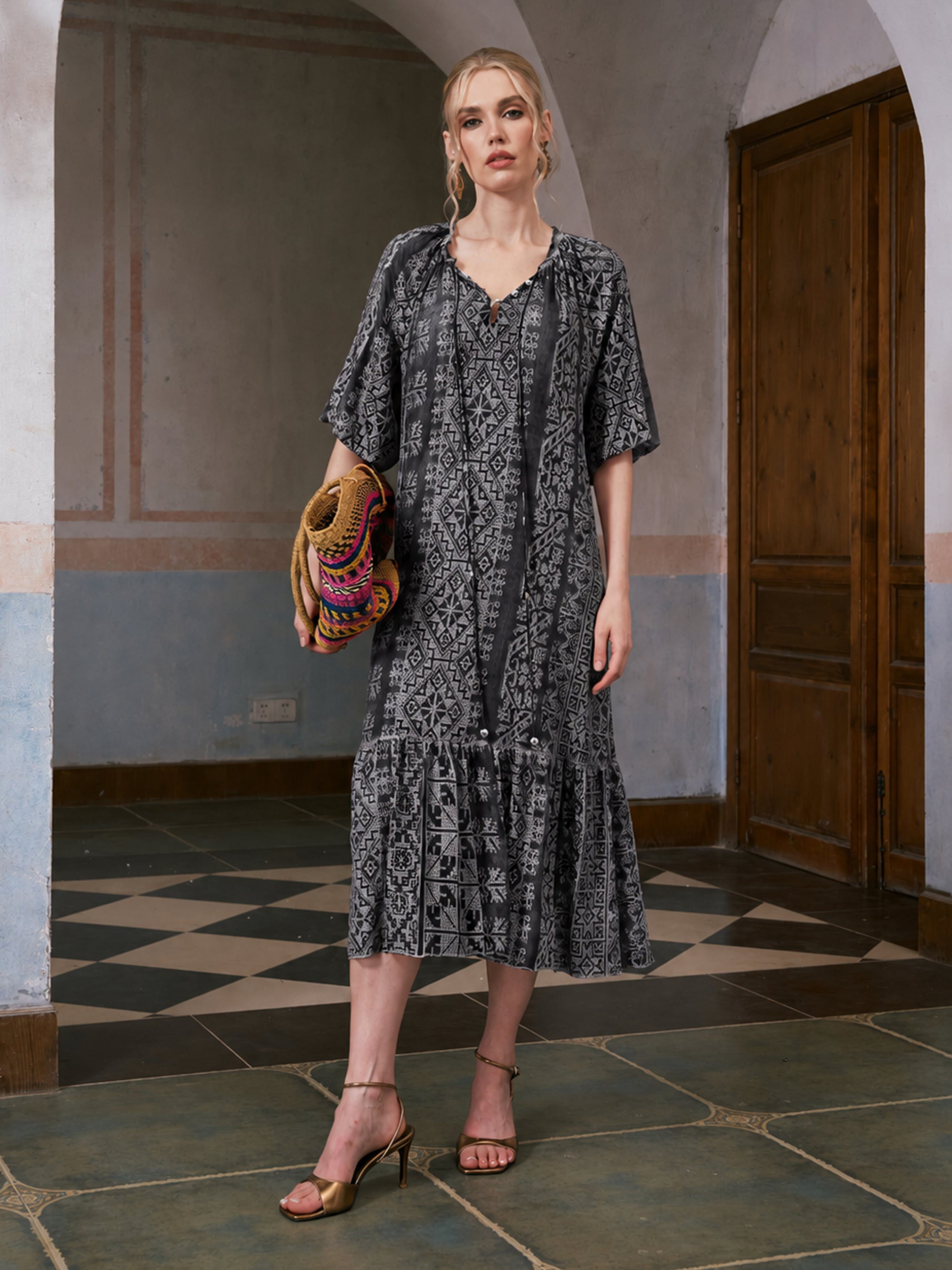 A woman stands indoors on a checkered tile floor with arched walls in the background, wearing the Social Queen Geometric Jacquard Mulberry Silk Dress. She holds a woven clutch and pairs the look with gold heels.