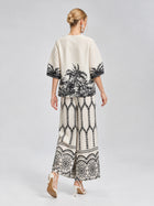 A woman stands with her back to the camera, wearing Social Queen’s Geometric Print Pleated Wide-Leg Pants in cream with a black geometric pattern, paired with white high-heeled sandals against a plain background.