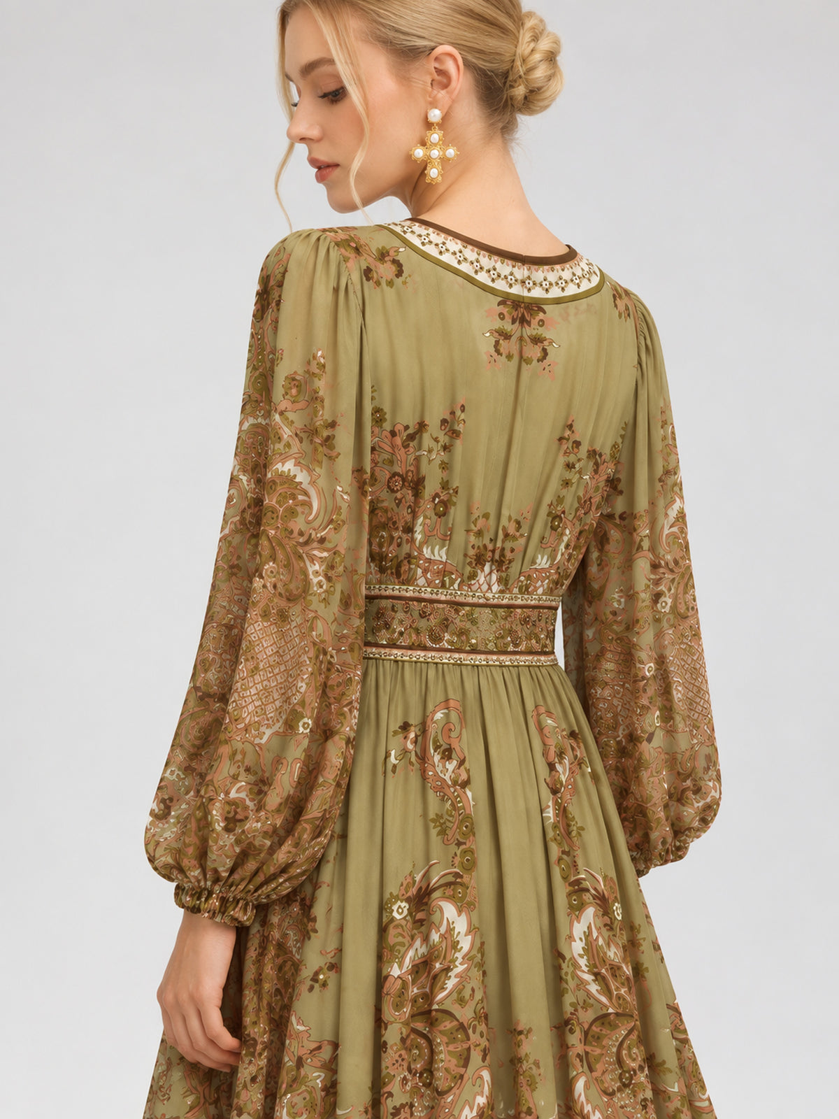 A woman wearing the Social Queen Paisley Print Mulberry Silk Dress—an olive green, long-sleeved dress adorned with a paisley floral print, puffed sleeves, and intricate detailing—poses against a plain background. This striking piece exemplifies effortless resort wear elegance.