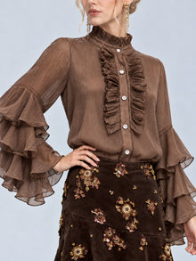 A woman wearing the Social Queen Pleated Layered Ruffle Blouse in brown, featuring tiered bell sleeves, paired with a dark brown floral embroidered skirt stands with one hand on her hip.