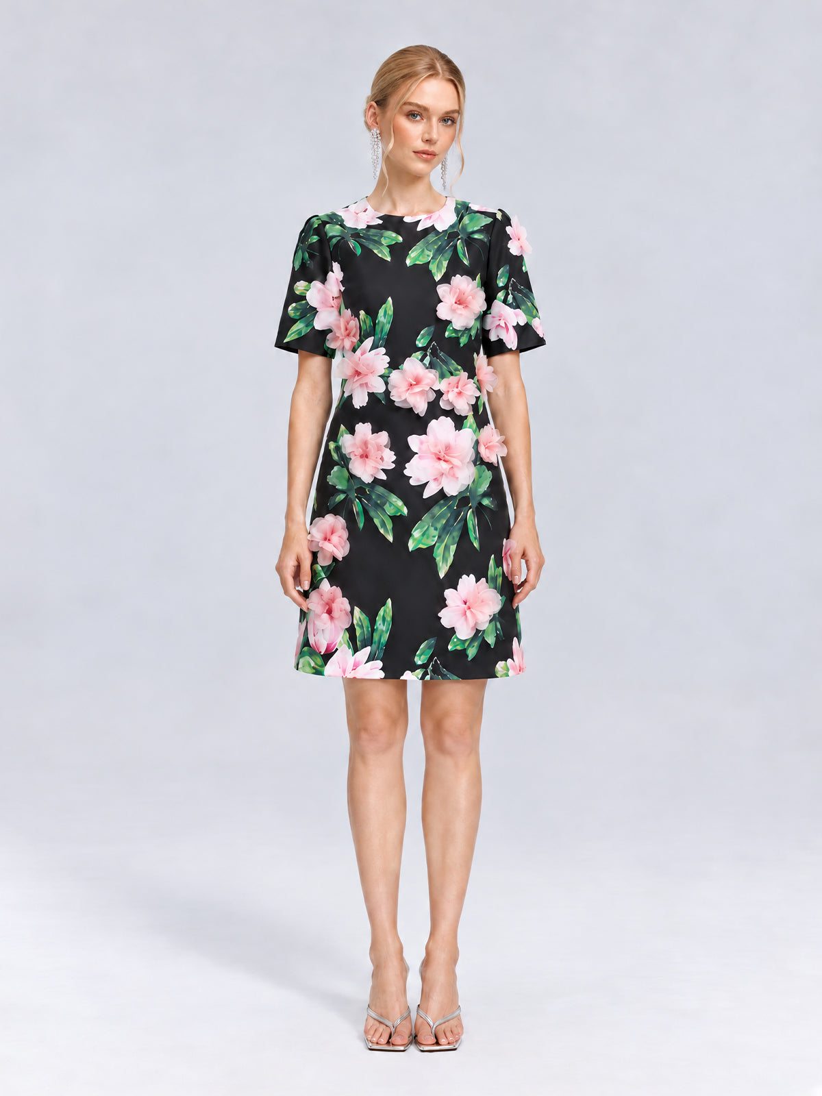 A woman stands against a light grey background wearing the Social Queen 3D Peony Floral Shift Dress, a short-sleeved, knee-length black shift dress adorned with soft pink peony flowers and green leaves, paired with open-toe heels.