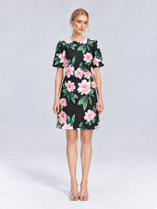 A woman stands against a light grey background wearing the Social Queen 3D Peony Floral Shift Dress, a short-sleeved, knee-length black shift dress adorned with soft pink peony flowers and green leaves, paired with open-toe heels.