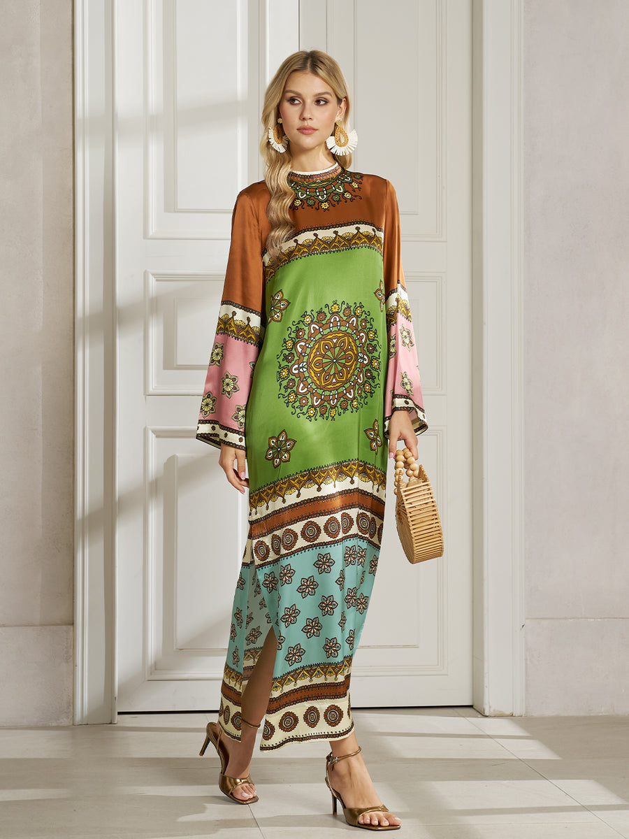 A woman stands indoors wearing a long, colorful patterned dress with wide sleeves, holding a woven handbag and wearing heeled sandals.