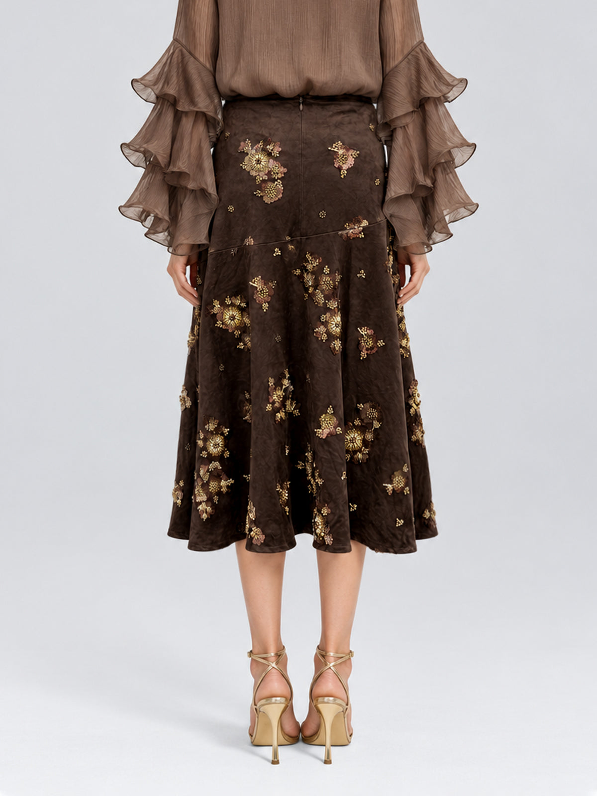 A person wearing a brown, long-sleeve blouse with layered ruffle cuffs and the Social Queen Floral Beaded Midi Skirt—a brown suede A-line skirt adorned with delicate floral embroidery—paired with beige high-heeled sandals, standing against a plain background.