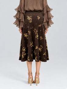 A person wearing a brown, long-sleeve blouse with layered ruffle cuffs and the Social Queen Floral Beaded Midi Skirt—a brown suede A-line skirt adorned with delicate floral embroidery—paired with beige high-heeled sandals, standing against a plain background.