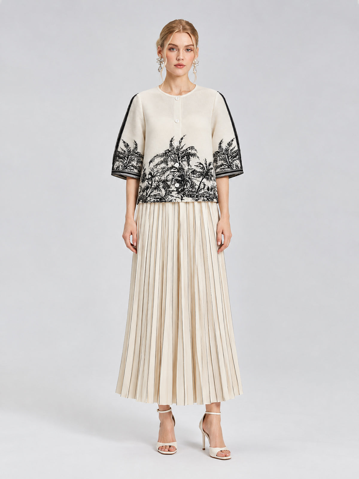 A woman stands against a plain light gray background wearing a cream top with black palm tree print and elbow-length sleeves, paired with Social Queen’s Striped Pleated Wide-Leg Pants and white heeled sandals.