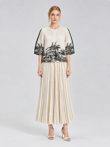A woman stands against a plain light gray background wearing a cream top with black palm tree print and elbow-length sleeves, paired with Social Queen’s Striped Pleated Wide-Leg Pants and white heeled sandals.