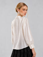A woman with blonde hair styled in an updo wears the Social Queen Floral Vine Embroidered Blouse—an ivory, shiny, long-sleeve top—paired with a black skirt in a relaxed straight silhouette. She is shown from the back, looking over her shoulder against a plain light background.