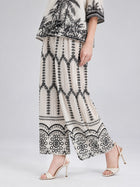 A person wearing the Social Queen Geometric Print Pleated Wide-Leg Pants, which feature a striking black geometric and floral pattern, paired with an off-white top adorned with black designs and white high-heeled sandals.