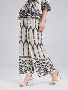 A person wearing the Social Queen Geometric Print Pleated Wide-Leg Pants, which feature a striking black geometric and floral pattern, paired with an off-white top adorned with black designs and white high-heeled sandals.
