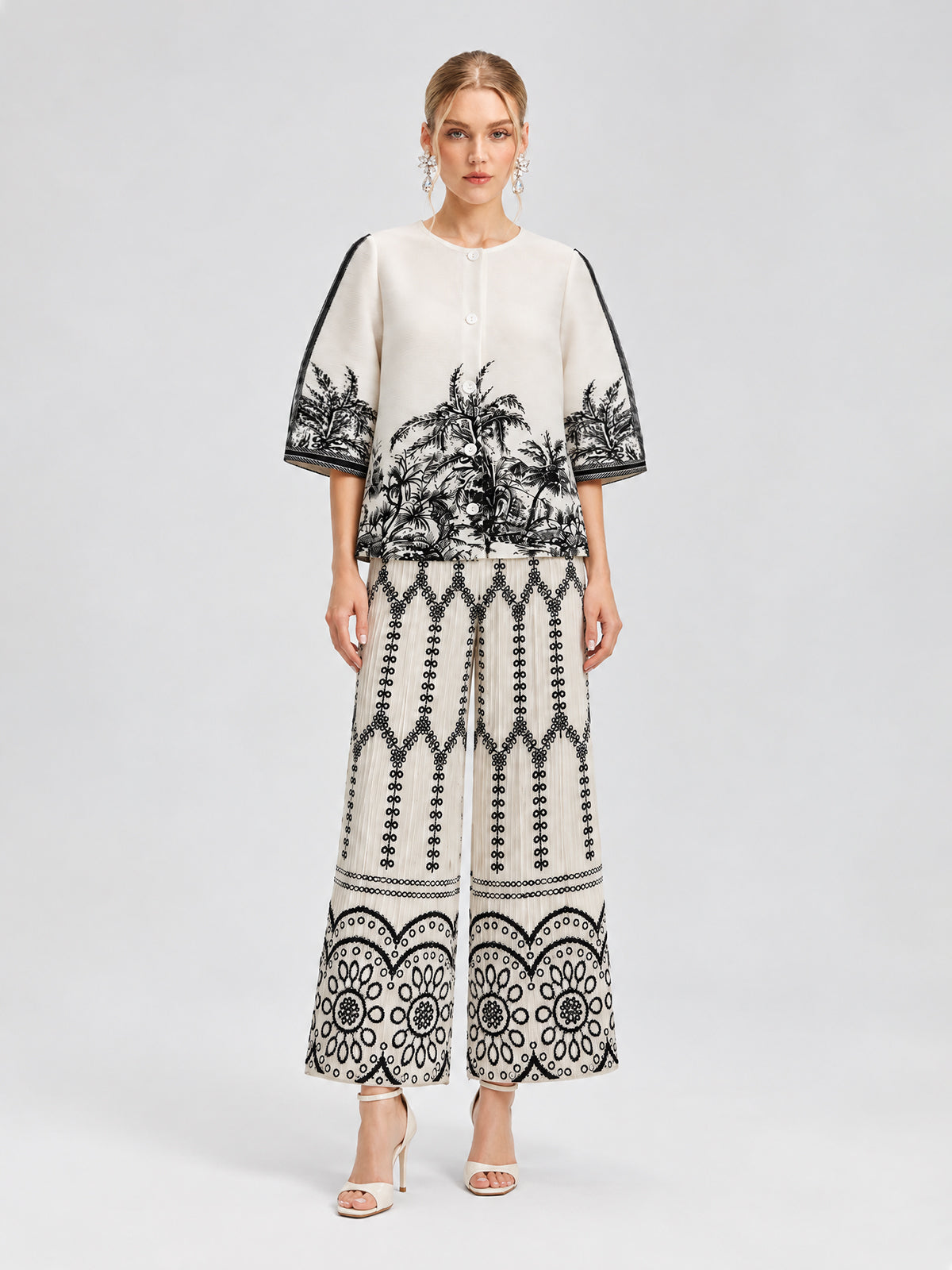 A woman stands against a neutral background wearing the Social Queen Palm Tree Print Pleated Blouse, a cropped, short-sleeve top adorned with black botanical and geometric patterns, paired with wide-leg pants featuring a palm tree print and open-toe heels.
