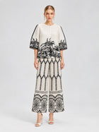 A woman stands against a neutral background wearing the Social Queen Palm Tree Print Pleated Blouse, a cropped, short-sleeve top adorned with black botanical and geometric patterns, paired with wide-leg pants featuring a palm tree print and open-toe heels.