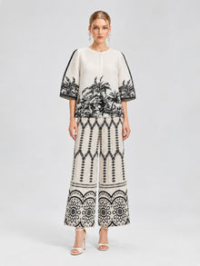 A woman stands against a neutral background wearing the Social Queen Palm Tree Print Pleated Blouse, a cropped, short-sleeve top adorned with black botanical and geometric patterns, paired with wide-leg pants featuring a palm tree print and open-toe heels.