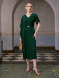 A woman wearing the Social Queen Asymmetric Fringe Midi Dress (belt included) in dark green, styled with gold heels and a woven handbag, stands indoors on a checkered tile floor with arched backgrounds.
