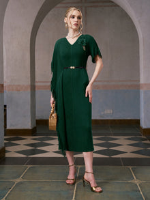 A woman wearing the Social Queen Asymmetric Fringe Midi Dress (belt included) in dark green, styled with gold heels and a woven handbag, stands indoors on a checkered tile floor with arched backgrounds.