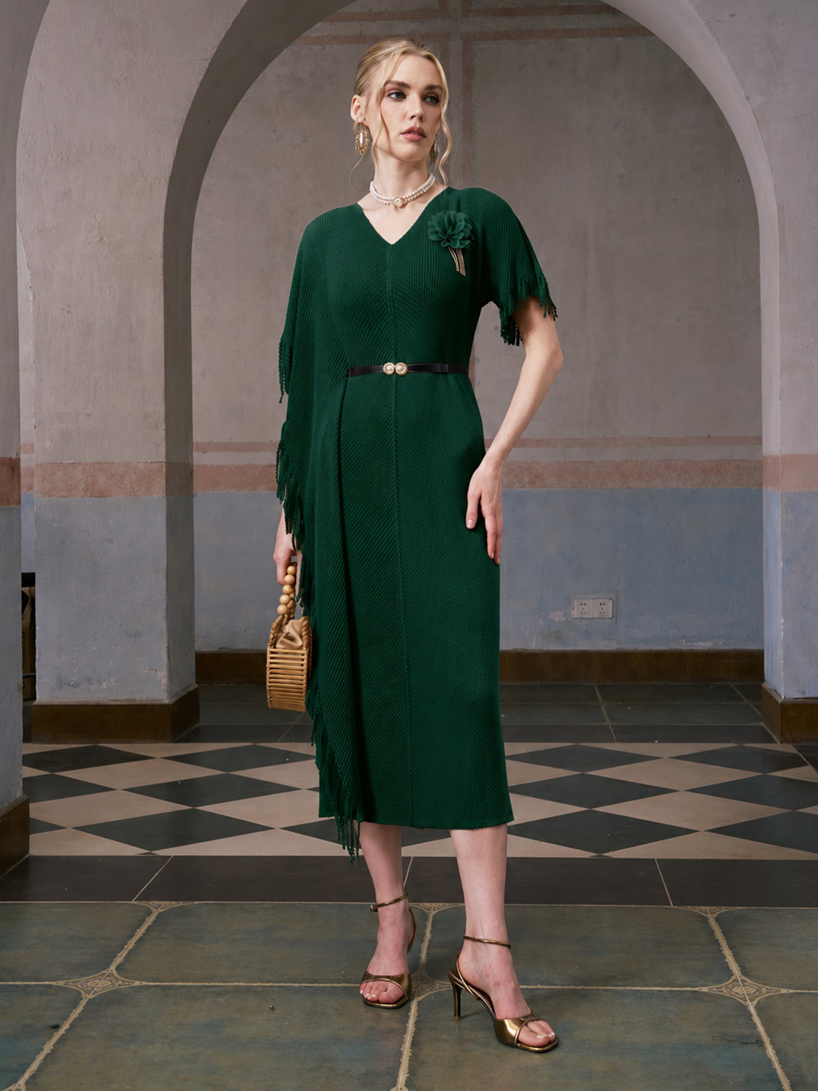 A woman wearing the Social Queen Asymmetric Fringe Midi Dress (belt included) in dark green, styled with gold heels and a woven handbag, stands indoors on a checkered tile floor with arched backgrounds.
